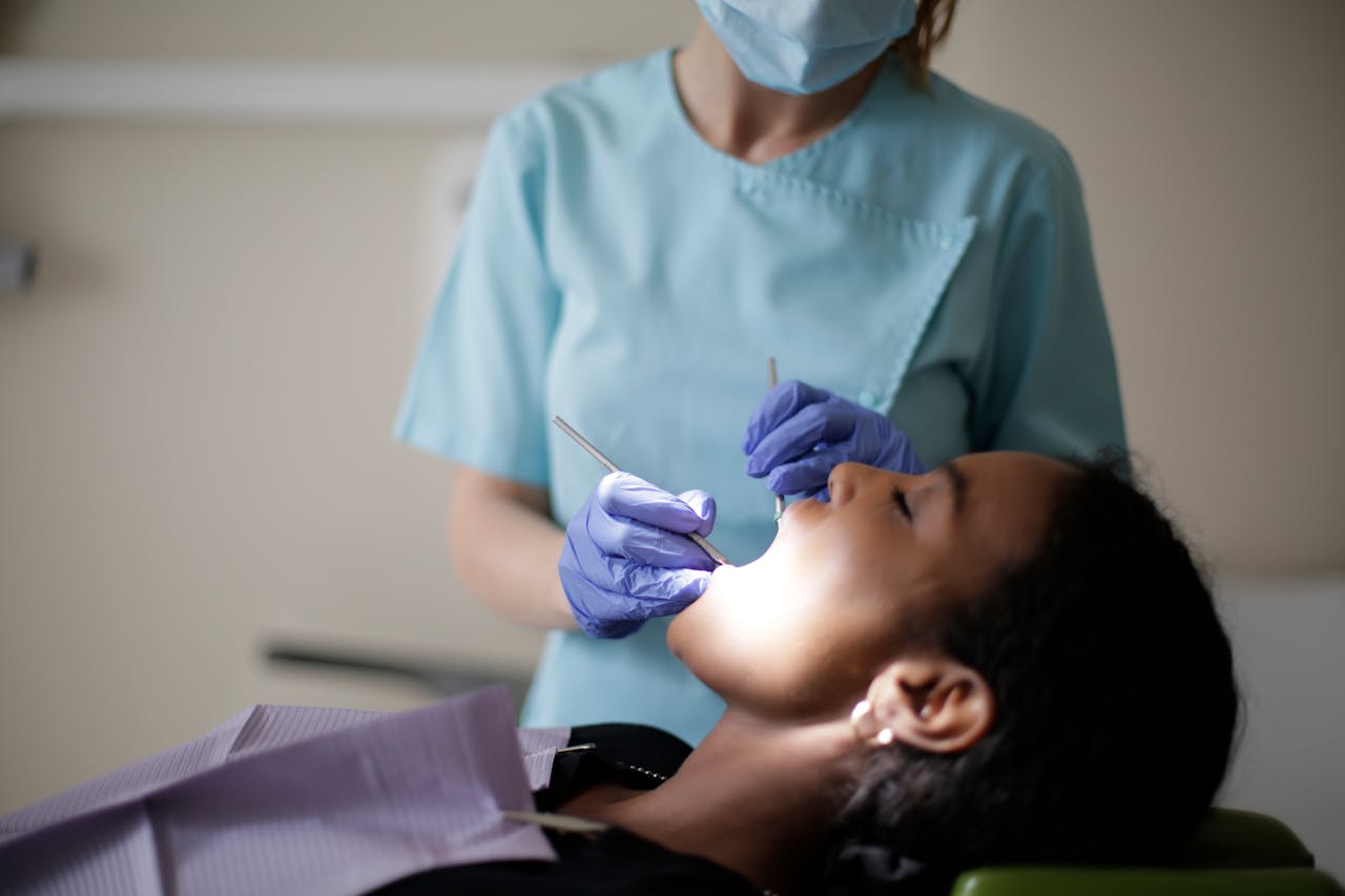 Focused female dentist in mask uniform and latex gloves using instruments for examining teeth of African American lady while patient lying with opened mouth and closed eyes