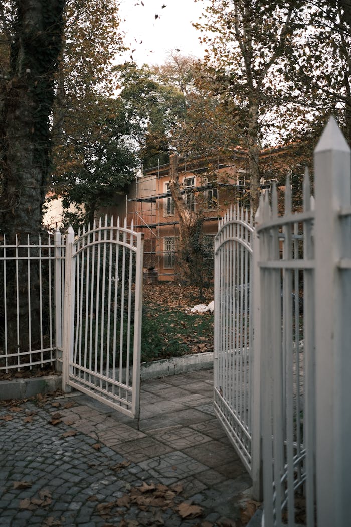 services-04 White metal gates open to a house surrounded by autumn trees and fallen leaves.