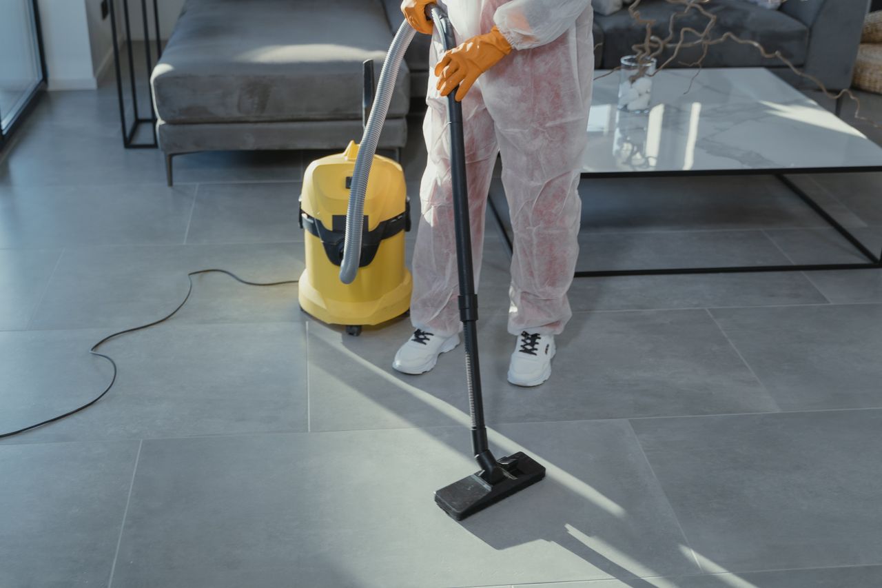 services-01 A professional cleaner in protective gear vacuuming the floor in a modern living room.