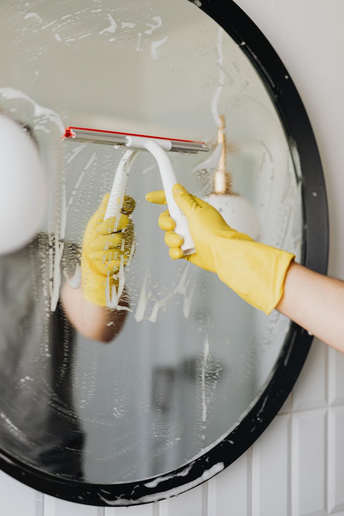 team-06 Faceless housekeeper in yellow rubber gloves cleaning round mirror from foam by squeegee in bathroom