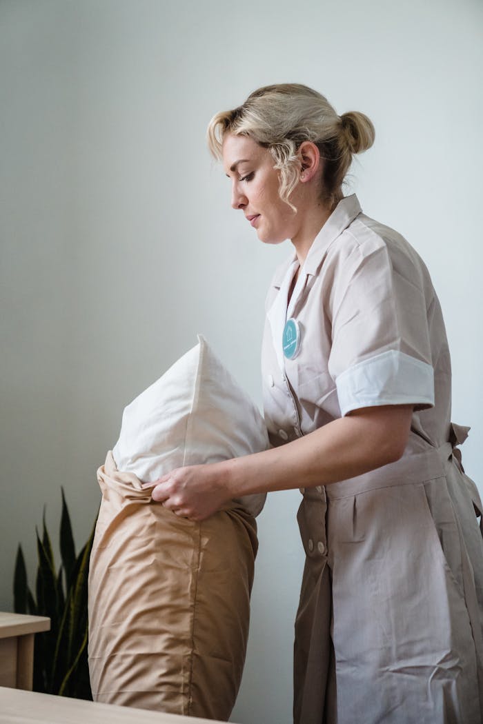 services-04 Hotel maid in uniform arranging pillows in a neatly styled guest room.