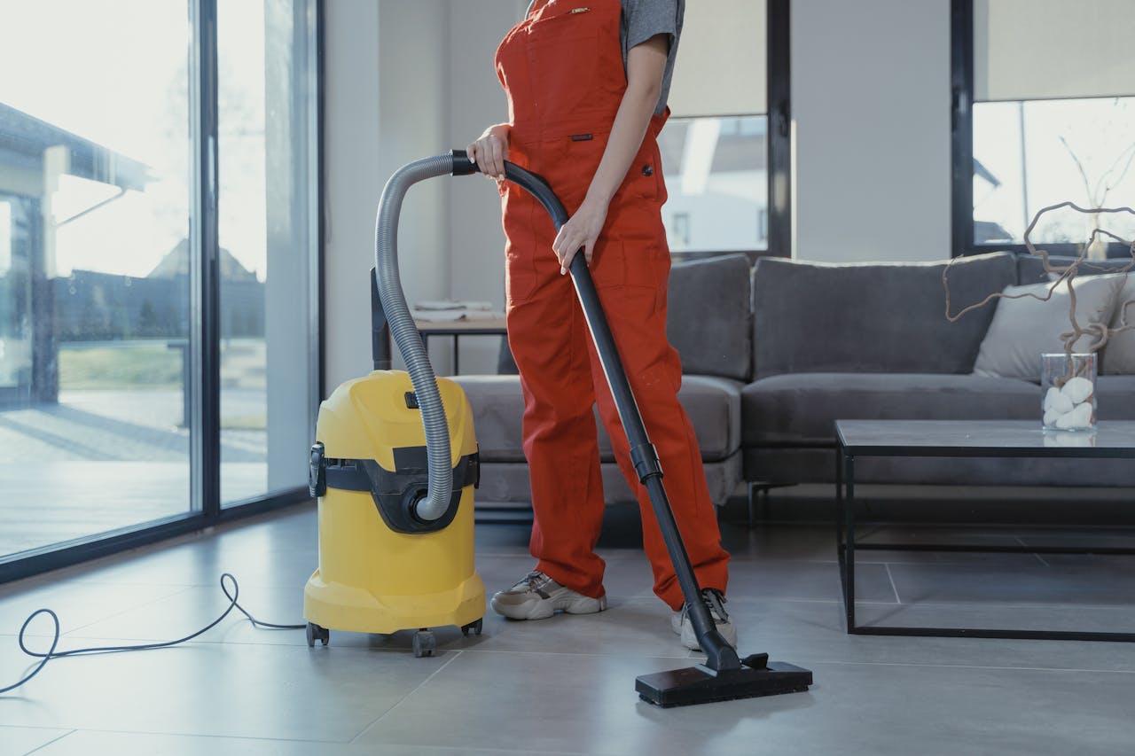 services-03 A professional cleaner in red coveralls vacuuming a modern living room with a yellow vacuum cleaner.