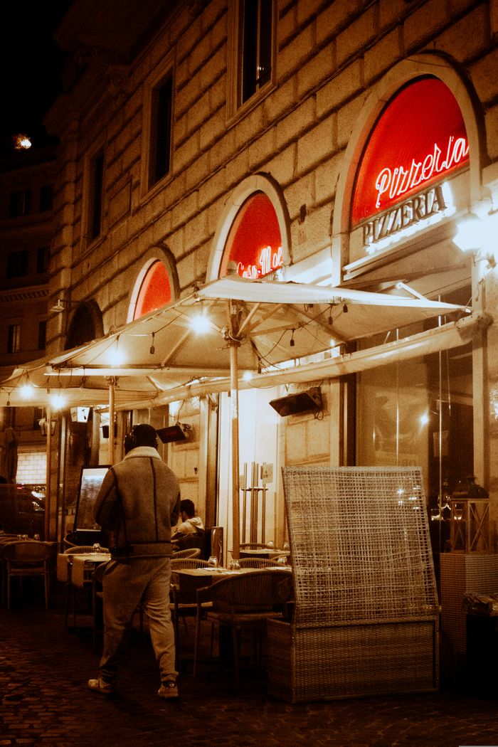 services-02 Cozy Italian pizzeria with outdoor dining under umbrellas on a bustling city night.